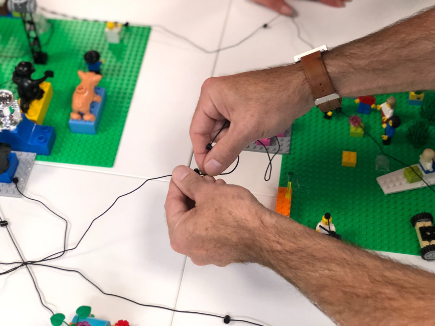 Manos conectando con cuerdas varios modelos LEGO sobre placas verdes para mostrar relaciones entre ideas